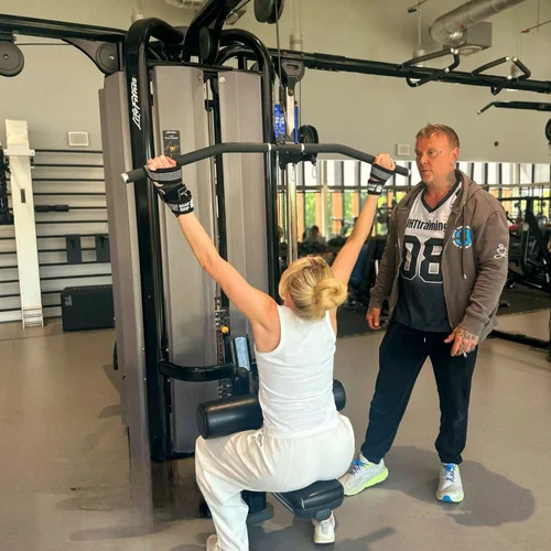 A woman lifting weights on a seated pulley machine with a trainer supervising in a gym.