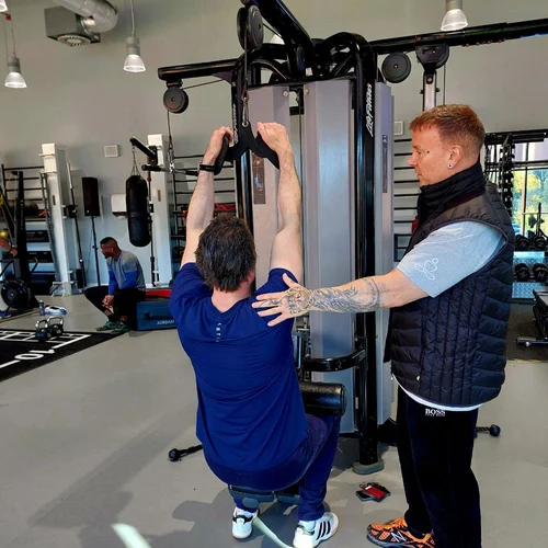 Person performing a seated lat pulldown exercise at a gym, guided by a trainer.